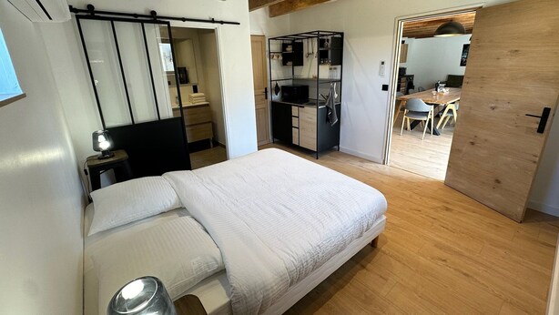 A bedroom with a bed, a bedside lamp, a wardrobe, and a kitchenette in the background. - Gîte de Condeiss (Condeissiat)