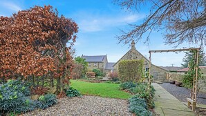 A garden with a stone pathway, a pergola, and a stone house with a sloped roof.