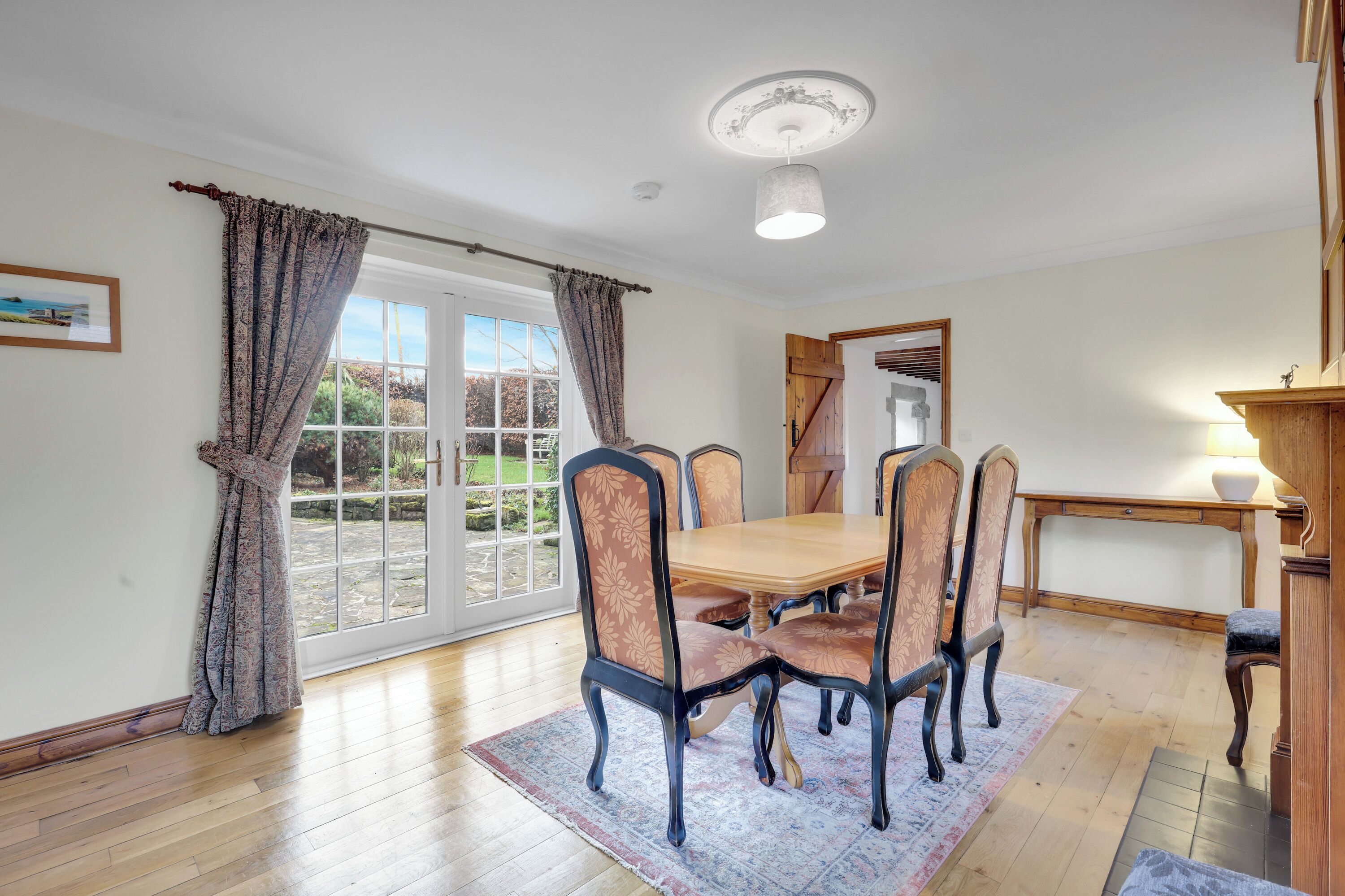 A dining area with a wooden table and chairs, a rug, and a view through a glass door to an outdoor garden.