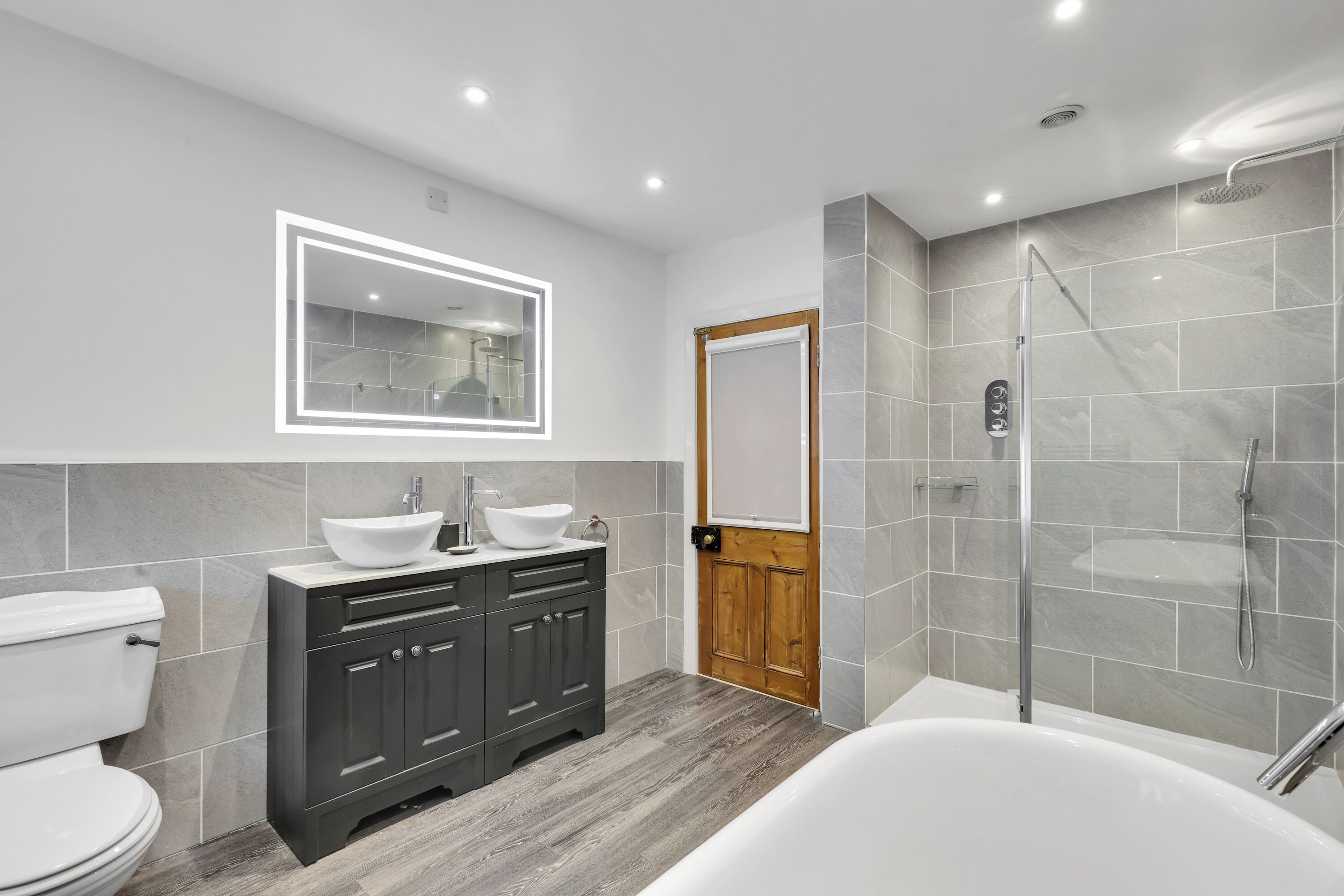 A modern bathroom with a white toilet, dark wooden cabinets, a large mirror with LED lighting, a white sink, and a walk-in shower.
