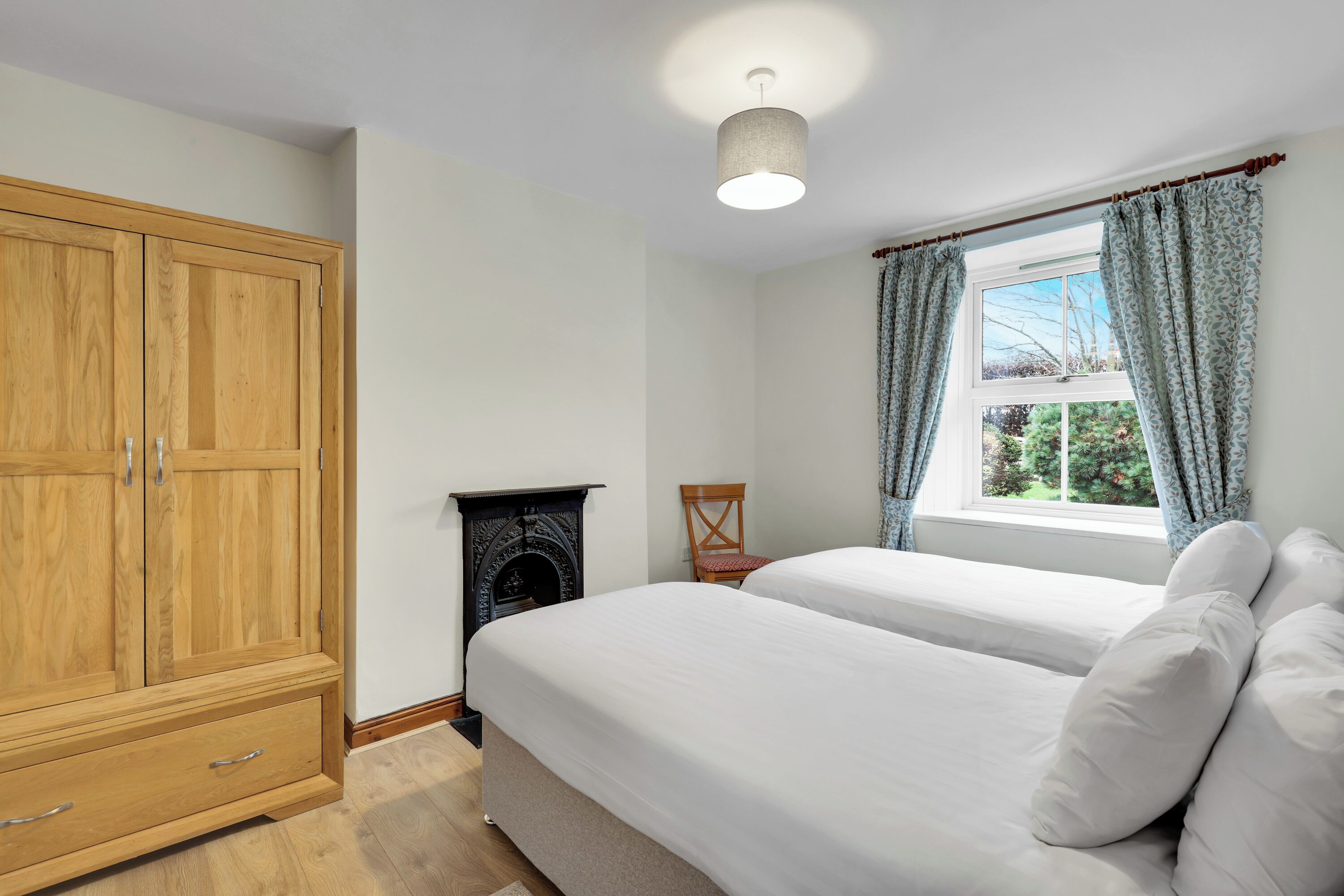 A bedroom with two beds, a wooden wardrobe, a fireplace, and a window with curtains.