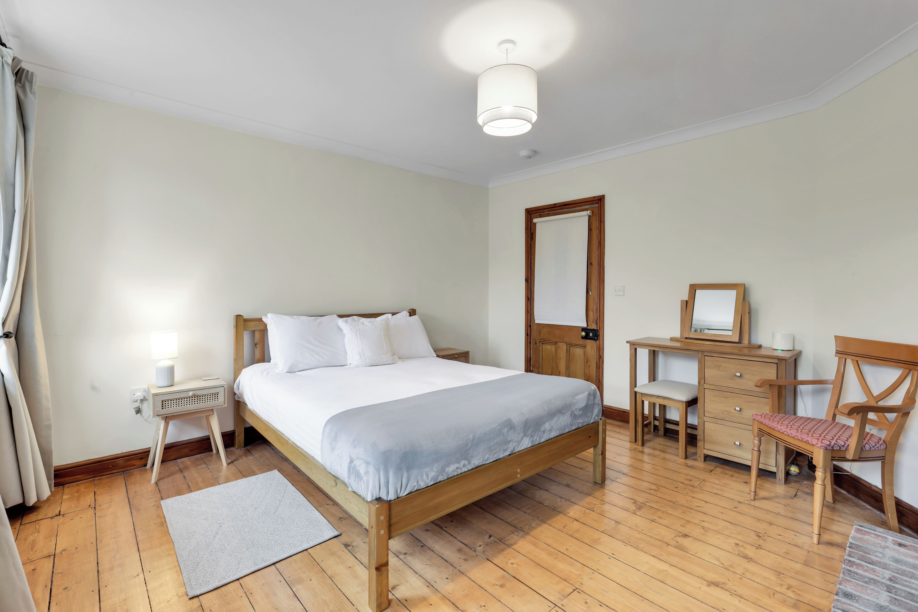 Un dormitorio con una cama de madera, una mesita de noche con lámpara, un aparador, una silla y un espejo.