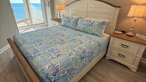 Une chambre à coucher comprenant un lit, une table de chevet, une lampe, un ventilateur de plafond et une vue sur l’océan, visible à travers une porte coulissante.