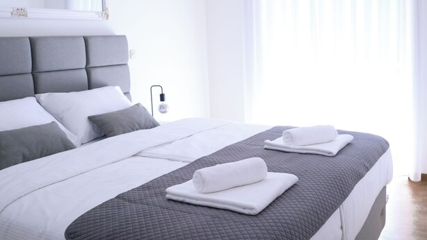 Ein modernes Hotelzimmer mit einem großen Bett, grau-weißer Bettwäsche und einer Nachttischlampe.