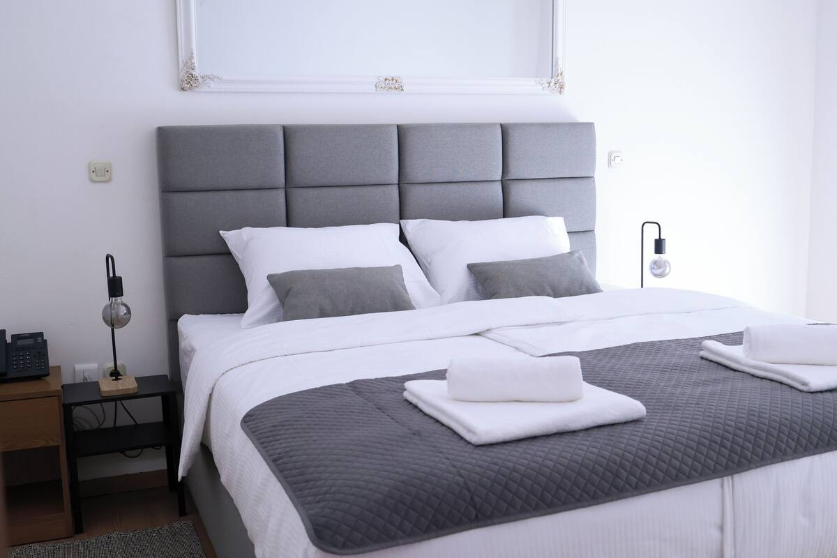 Un lit bien fait, avec une tête de lit grise, une literie blanche et un couvre-lit matelassé gris. On y trouve une table de chevet avec une lampe et une prise de courant.