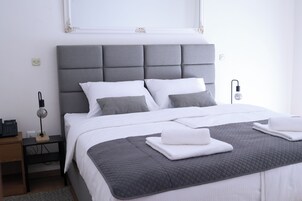 A neatly made bed with a grey headboard, white bedding, and a grey quilted throw. There is a bedside table with a lamp and a power outlet.