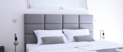 Una cama bien hecha con un cabecero gris, almohadas blancas y una colcha gris acolchada. Se ve una lámpara de noche y una mesita con una lámpara.