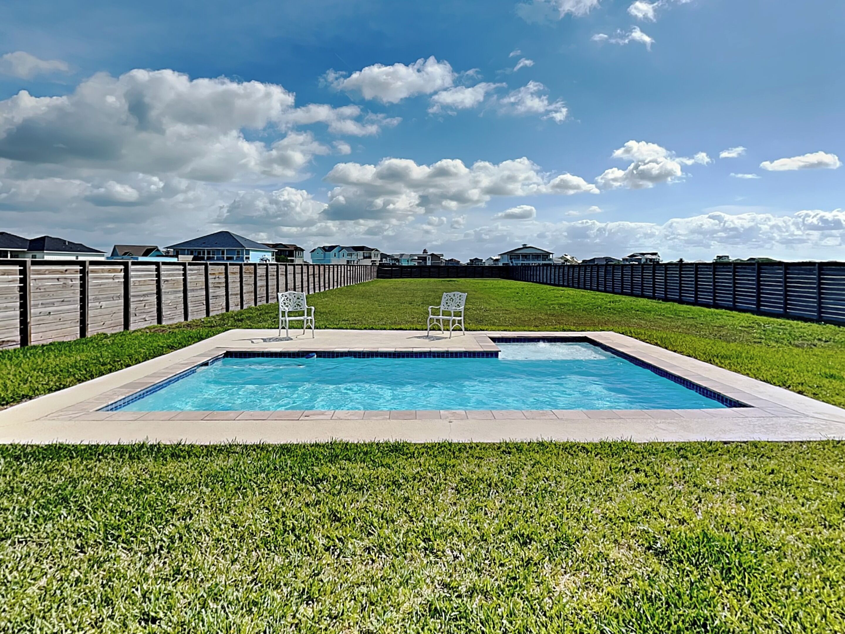 A rectangular swimming pool with two white chairs, surrounded by a wooden fence, with houses in the background.