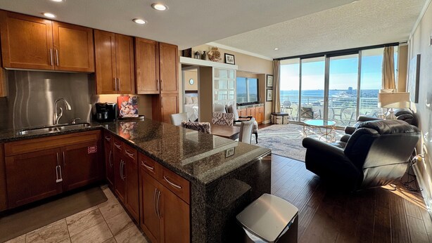 A modern kitchen with granite countertops, wooden cabinets, and a stainless steel sink. There is a dining area with a view of the ocean. - Beautiful Sunset and Gulf Views-2 bedroom / 2 bathroom (Biloxi)