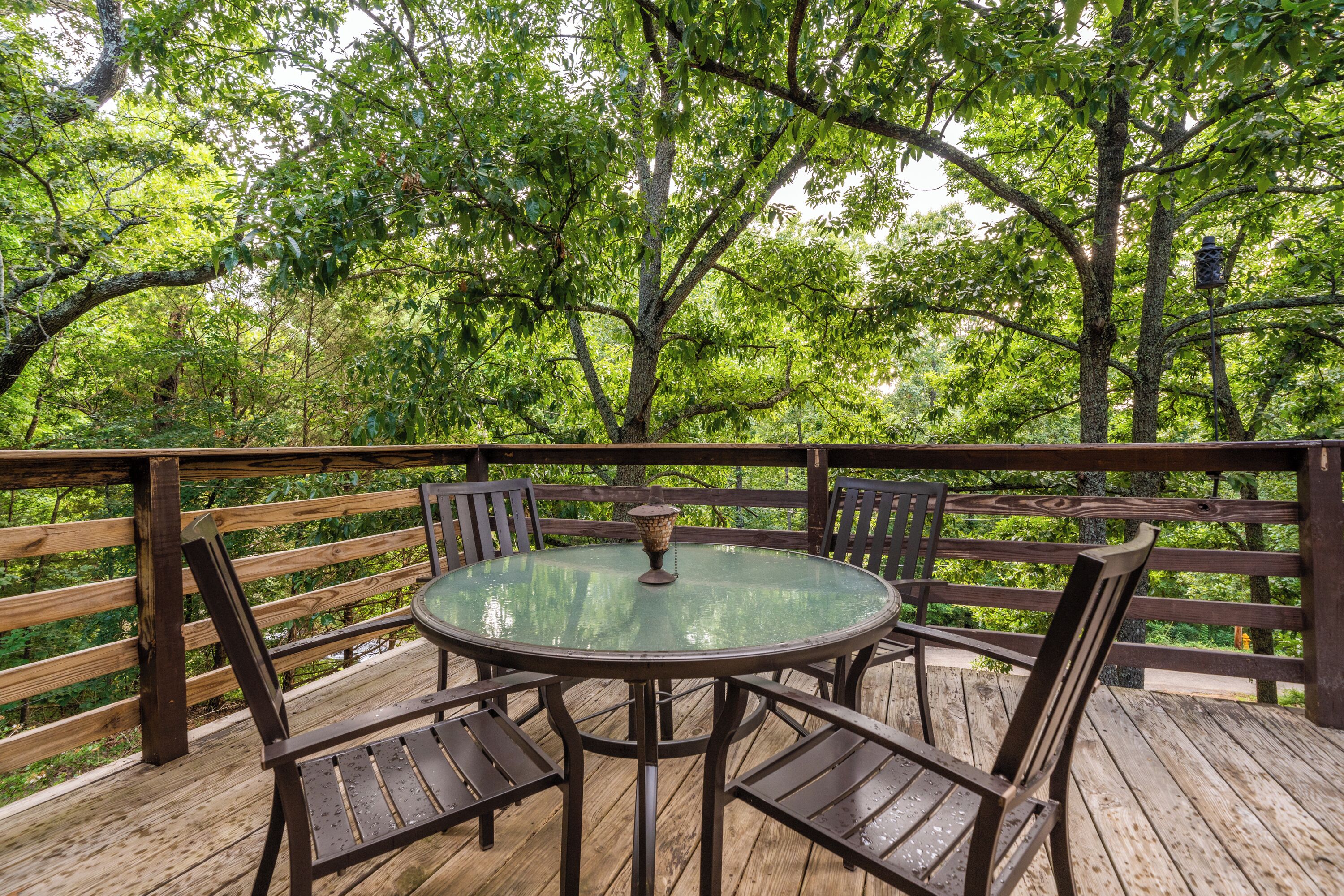 Een houten terras met een glazen tafel en stoelen, omringd door bomen.