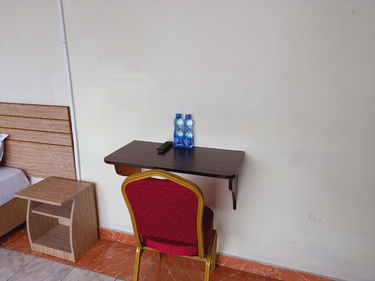 Una habitación pequeña con una cama de madera, una mesita de madera con dos botellas de agua y un mando a distancia, y una silla roja con estructura dorada.