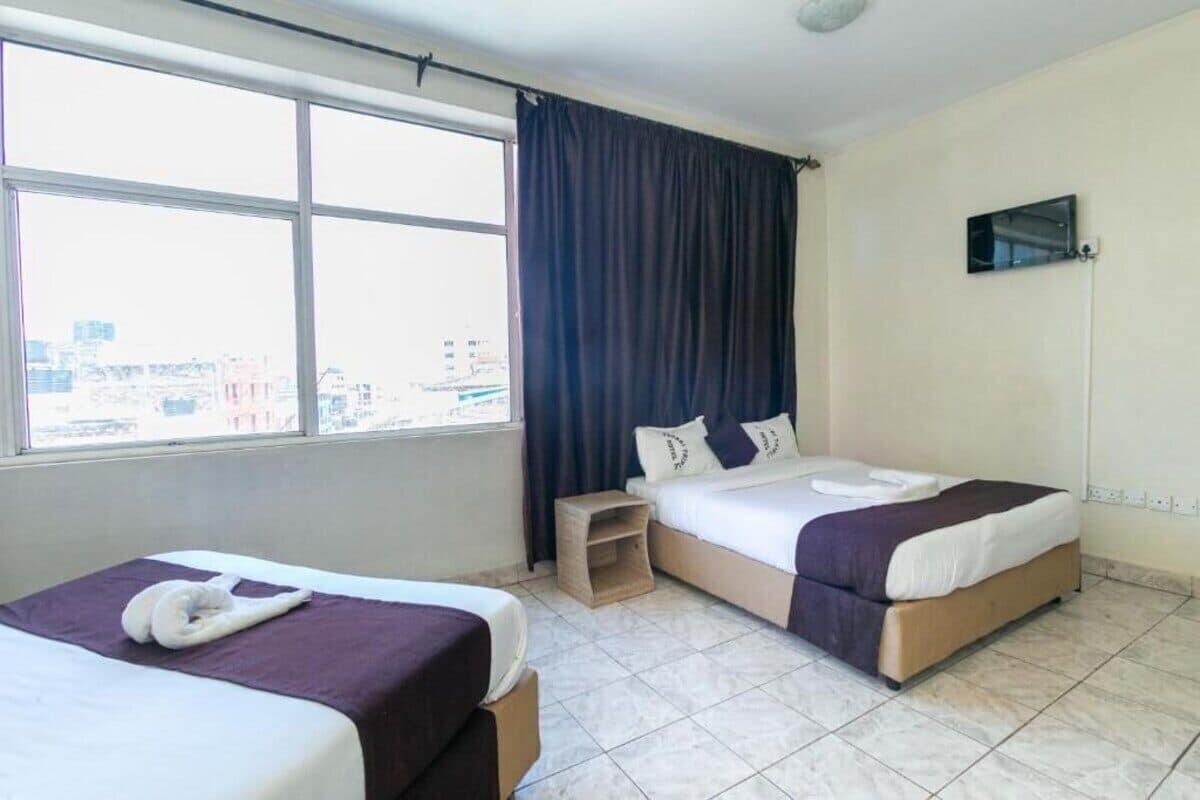 Habitación de hotel con dos camas, una mesita de noche de madera, televisor montado en la pared y un ventanal con cortinas.