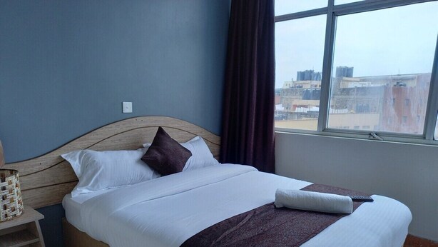 A neatly made bed in a room with a large window, a bedside table, and a view of buildings outside. - Nairobi Fahari Tripple Suites (Nairobi)