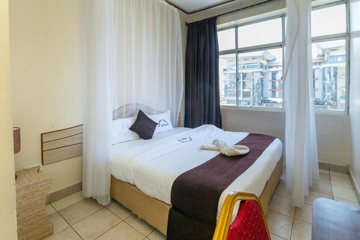 Una habitación de hotel con cama, ventana con cortinas, vista de edificios exteriores y un taburete de madera.