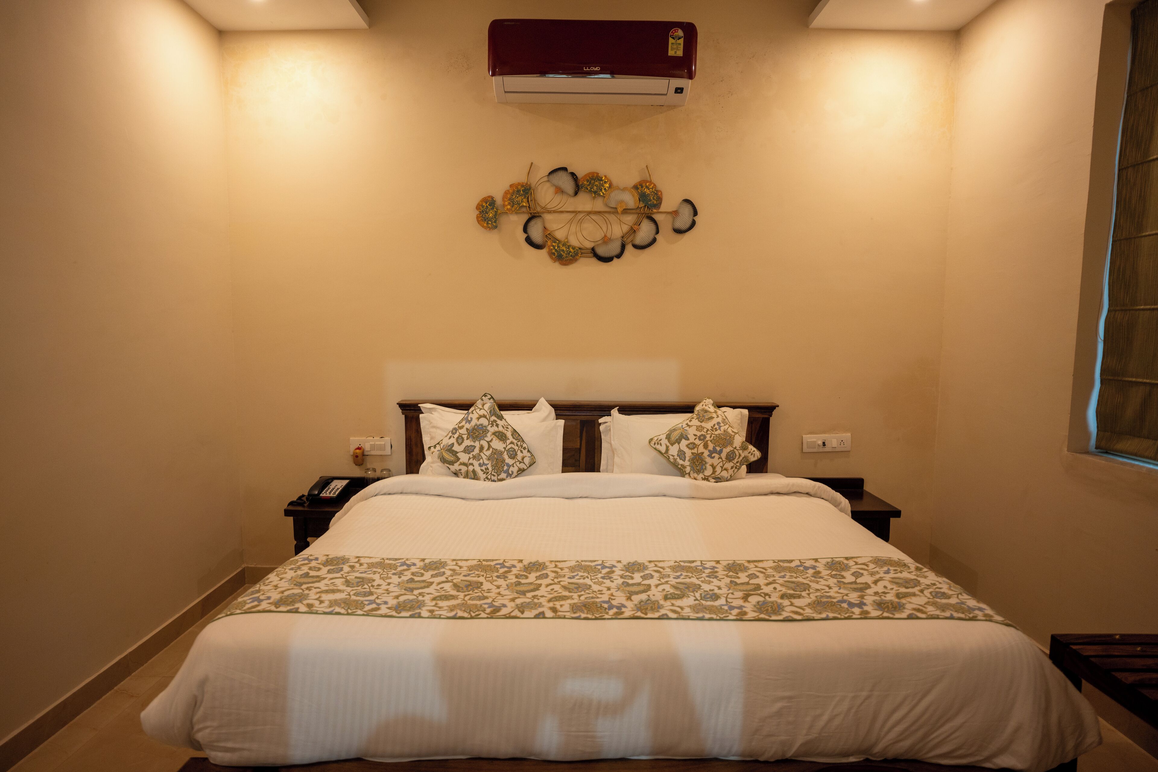 Un lit bien fait, recouvert d’une couvre-lit à motifs, un climatiseur fixé au mur et un élément décoratif mural au-dessus du lit.