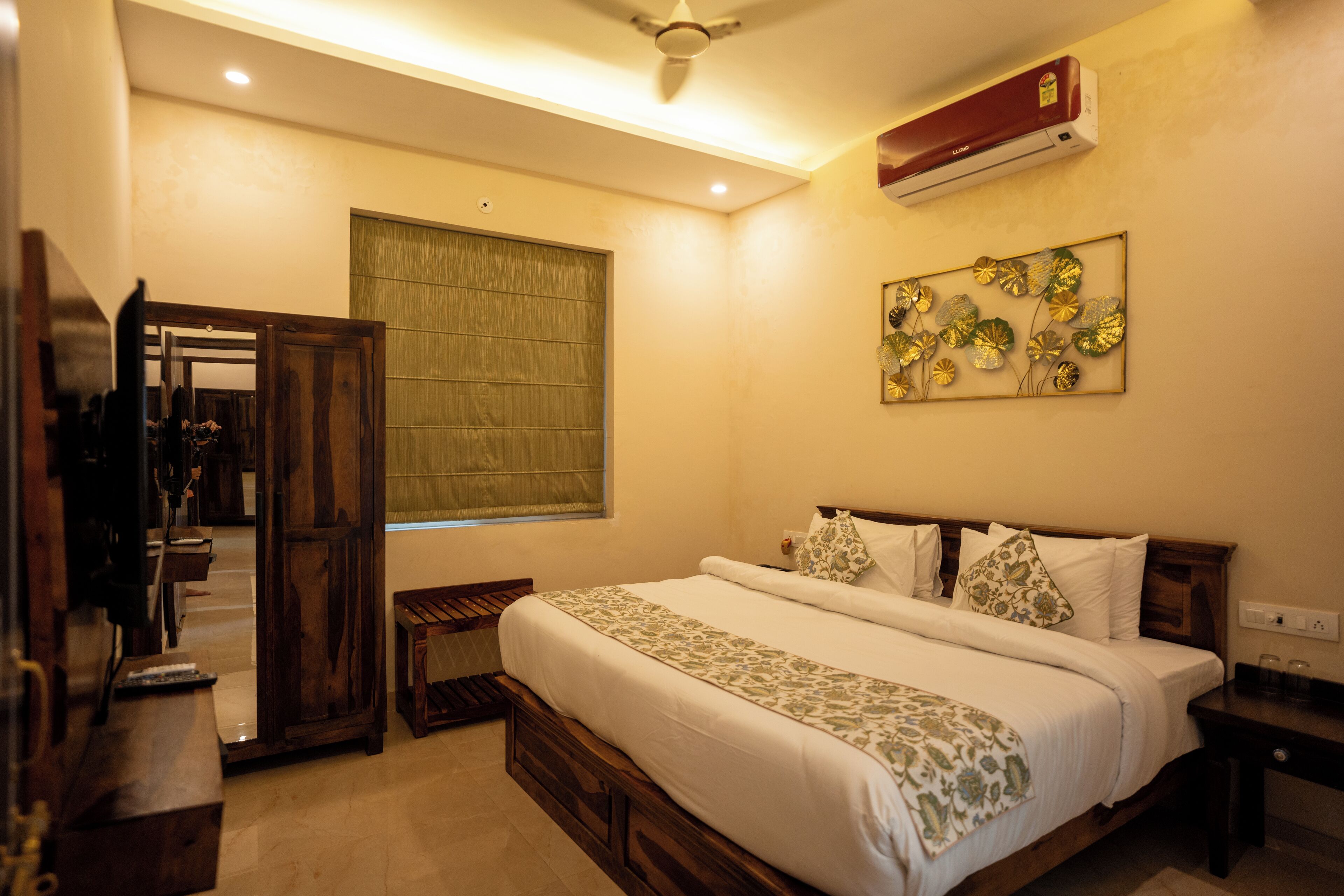 Une chambre d’hôtel avec un grand lit, une tête de lit en bois, une table de chevet, une petite table de chevet, un climatiseur mural, un ventilateur de plafond, une fenêtre avec un store vert et un élément décoratif mural.