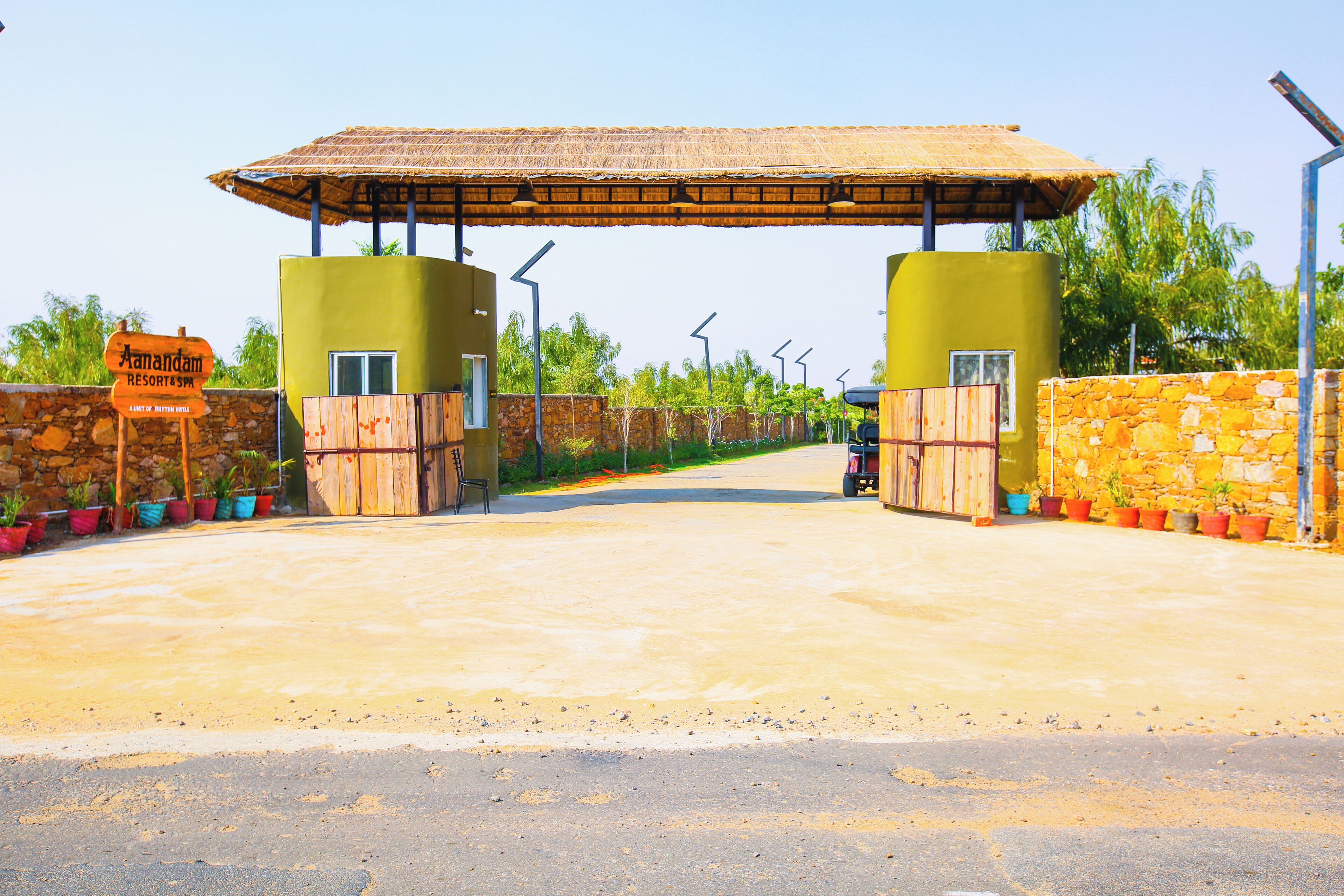 A resort entrance with a thatched roof, a sign indicating 'Anandam Resort', and a paved pathway leading inside.
