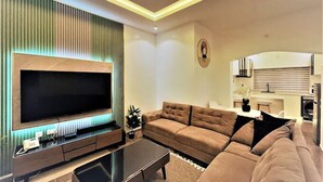 Ein modernes Wohnzimmer mit großem Flachbildfernseher, einer Couch, einem Couchtisch und einer Küchenzeile mit Spüle und Herd.