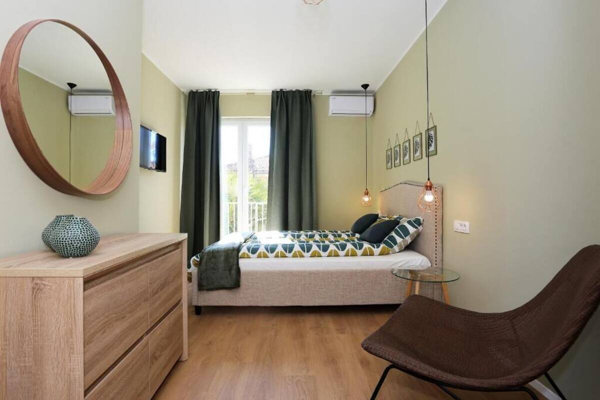 Une chambre à coucher avec un lit, une commode en bois, un miroir rond, une chaise et une petite table.