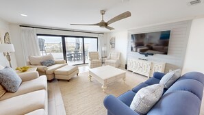 A spacious living room with a large flat-screen TV, a ceiling fan, and a balcony with outdoor furniture.