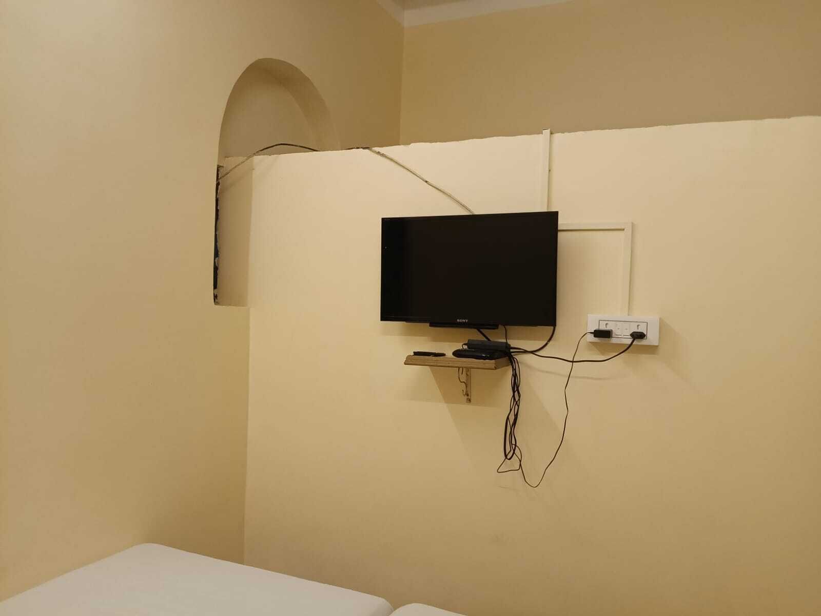 A room with a mounted television on the wall, a power outlet, and a small wooden shelf below it.