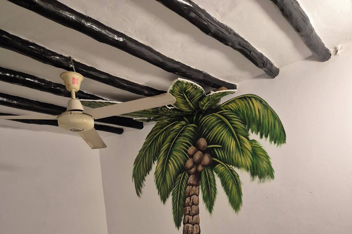 Un dormitori amb un llit, una petita taula de nit de fusta, un ventilador de sostre i un mural al mur amb una palmera.