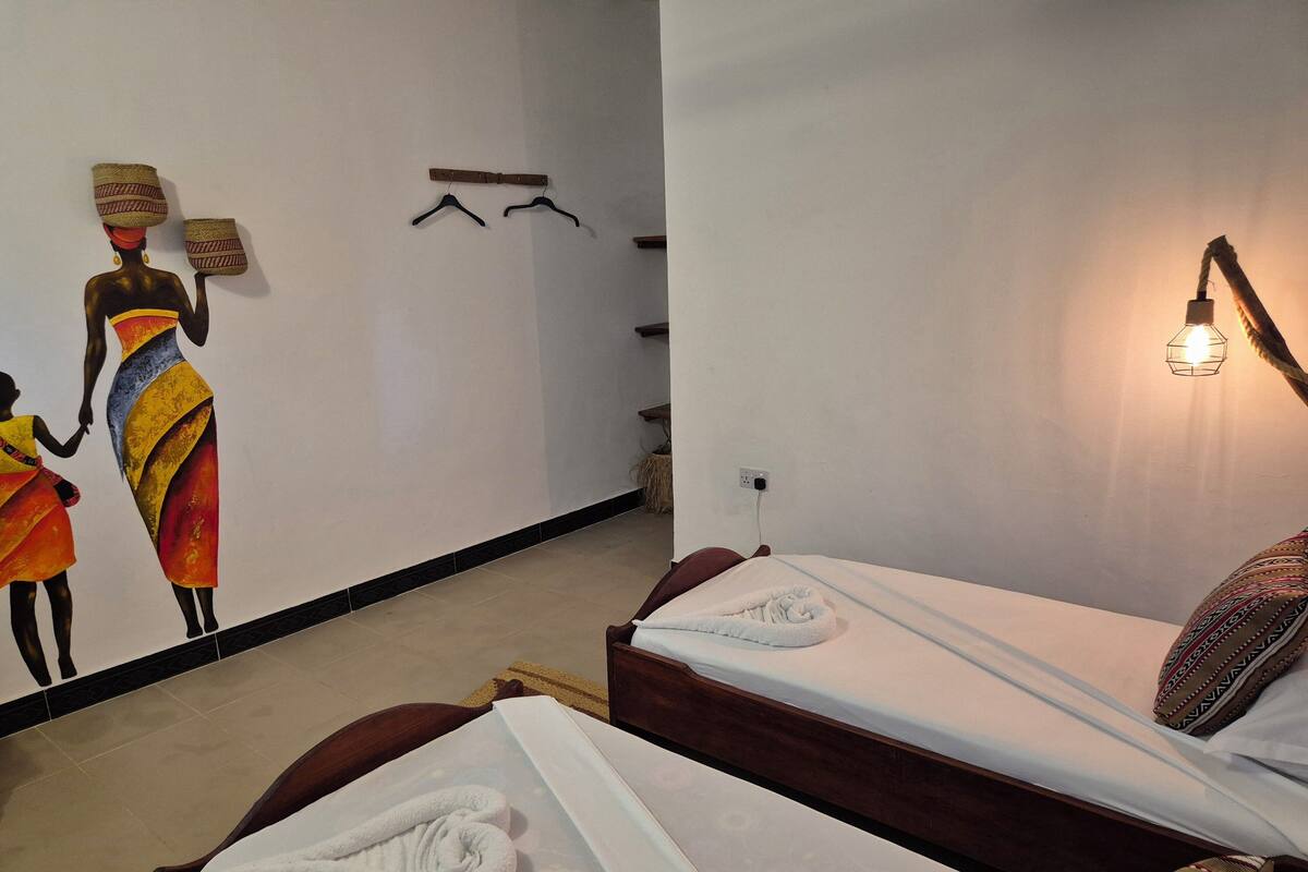 Habitació d'hotel amb un llit, una làmpada fixada a la paret, una obra d'art amb dues figures i una petita estanteria amb objectes penjats.