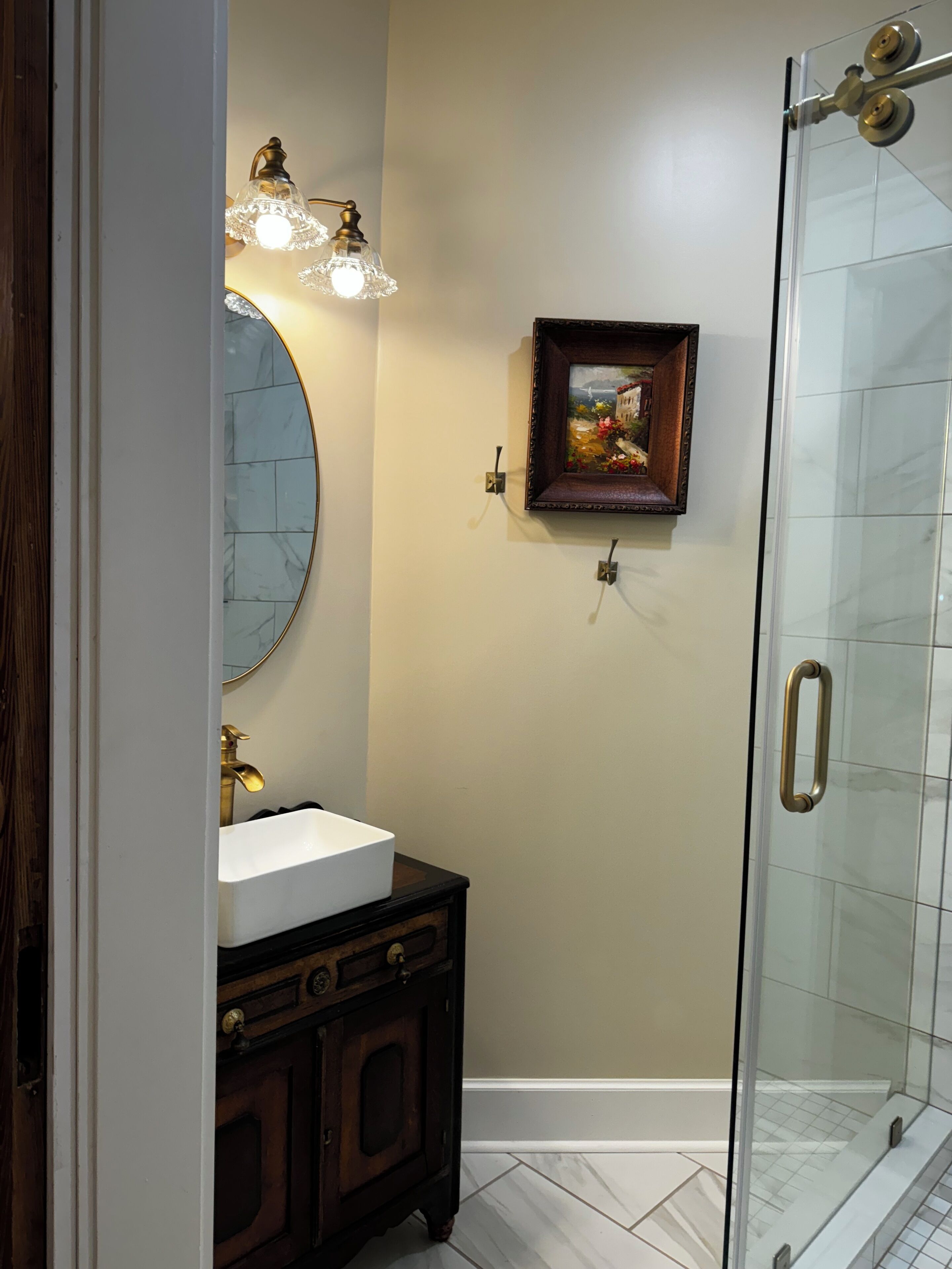 Une salle de bain avec une douche à parois de verre, un miroir rond, un lavabo avec un meuble en bois et un tableau encadré au mur.