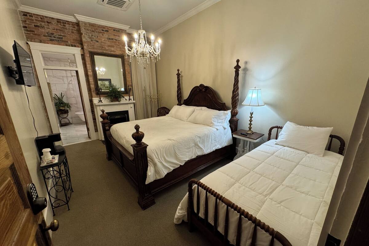 A bedroom with a large bed, a smaller bed, a chandelier, a fireplace, and a mirror.