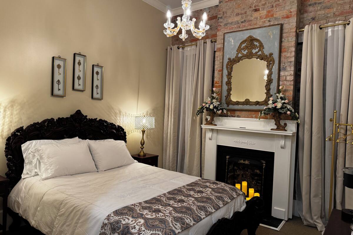 A bedroom with a large bed, a chandelier, a fireplace, and framed pictures on the wall.