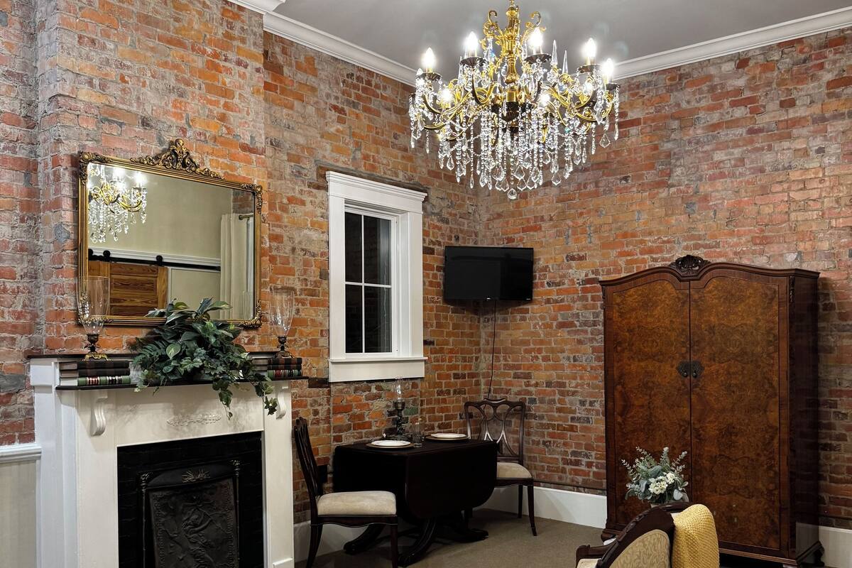 A room with brick walls, a chandelier, a fireplace, a dining table with chairs, a sofa, and a large wooden cabinet.