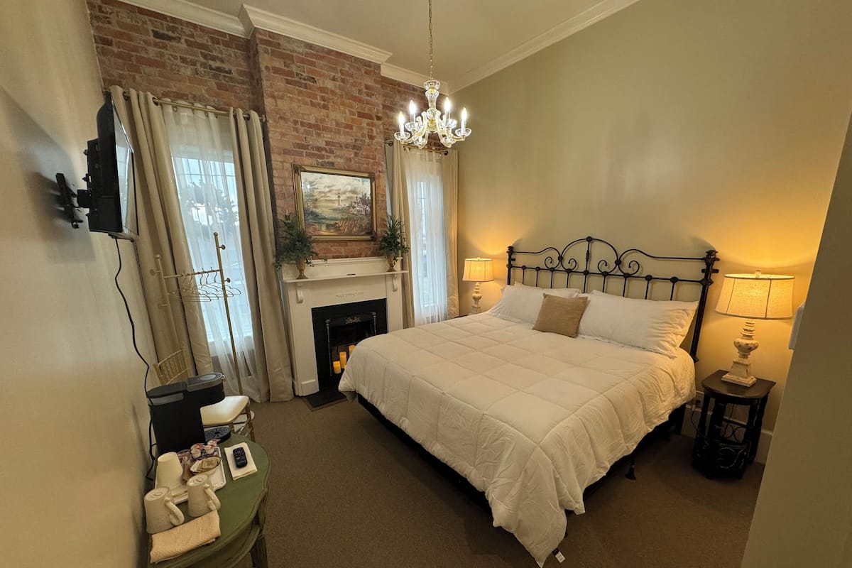 A bedroom with a large bed, a fireplace, a chandelier, and a painting on the wall.