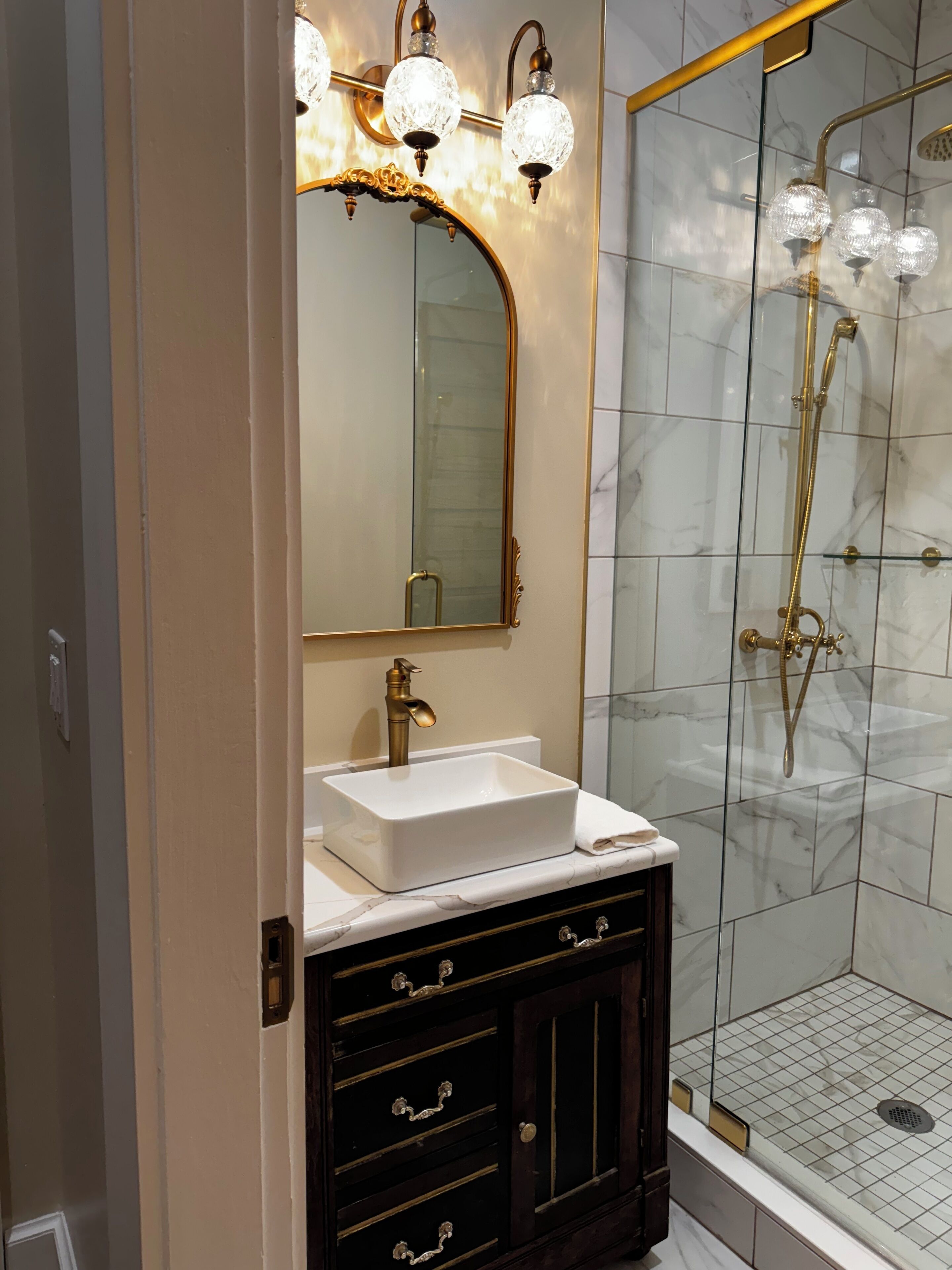 Un baño con lavamanos blanco, elementos dorados y una ducha con mampara de vidrio y detalles dorados.