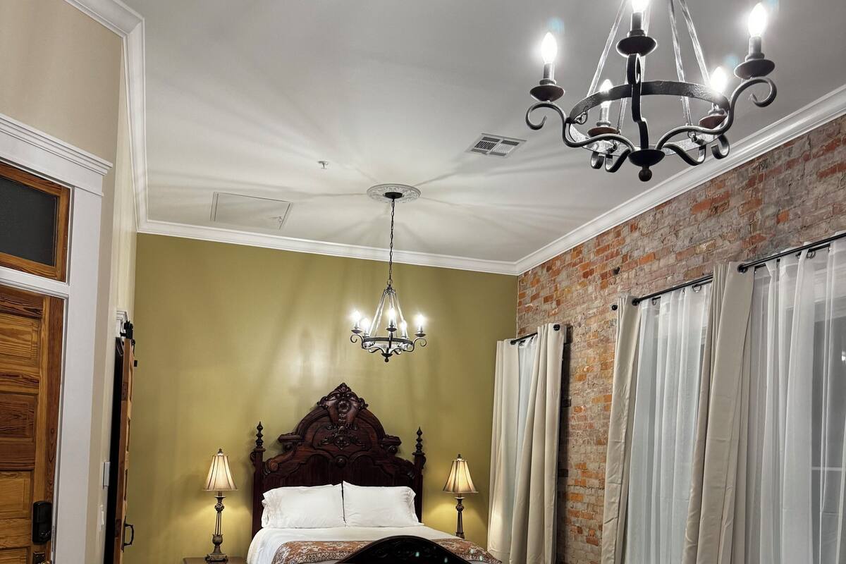 A hotel room with a large bed, two armchairs, a small table, a chandelier, and a brick wall.