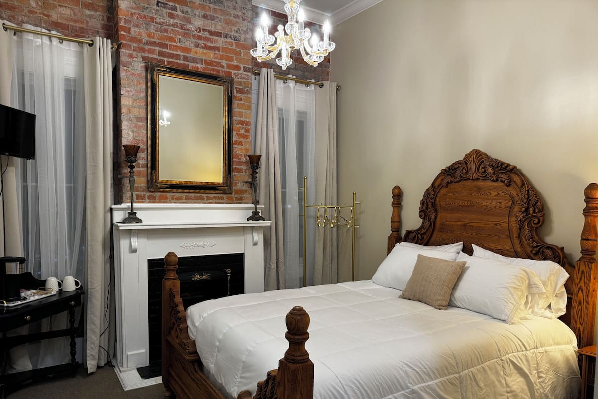 A bedroom with a brick fireplace, a wooden bed with a carved headboard, a chandelier, and a mirror above the fireplace.