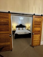 A bedroom with a bed, two bedside tables with lamps, a chandelier, and wooden doors.