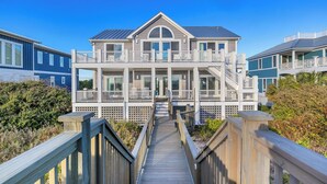 A multi-story beachfront house with a wooden deck leading to the entrance, surrounded by lush greenery. - One More Day Caswell: Oceanfront Home With Elevator (Oak Island)