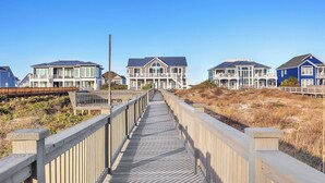 A wooden boardwalk leading to a row of beachfront houses with balconies, a clear sky, and dry grass. - One More Day Caswell: Oceanfront Home With Elevator (Oak Island)