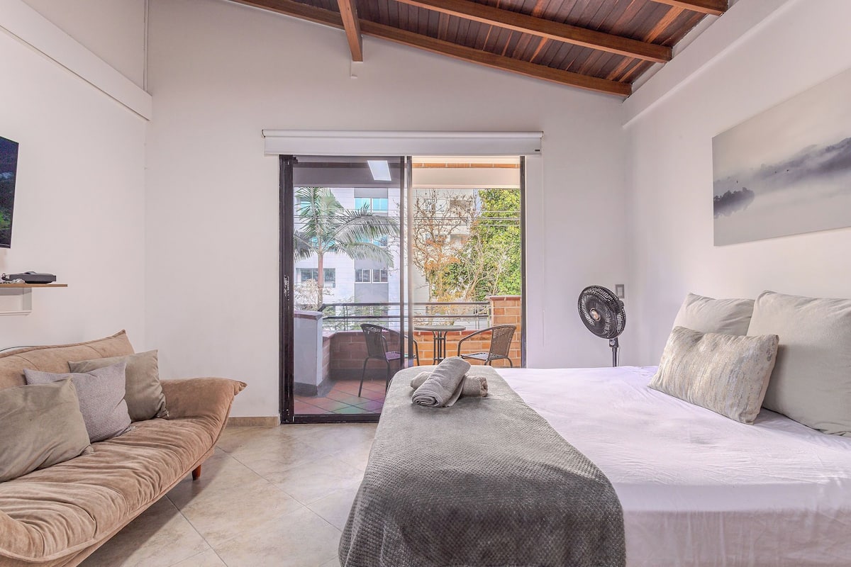 A bedroom with a bed, a sofa, a fan, and a sliding glass door leading to an outdoor area with a table and chairs.