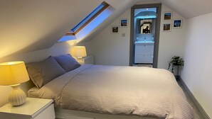 Ein Schlafzimmer mit Dachfenster, einem Bett mit Staufächern, Nachttischlampen, einem kleinen Badezimmer und gerahmten Bildern an der Wand.