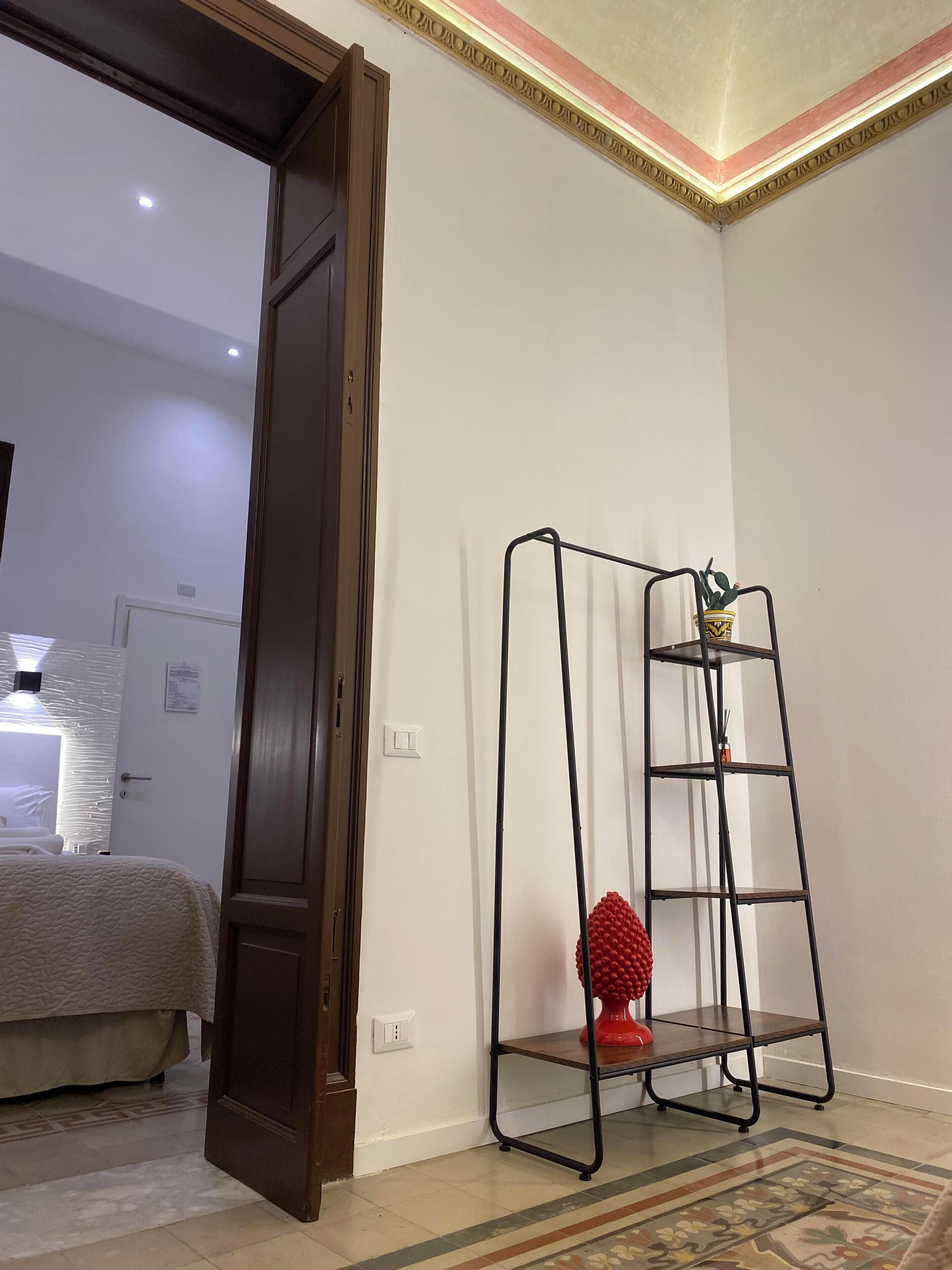Ein Hotelzimmer mit einem Bett, einer Holztür, einem schwarzen Metallregal mit einem roten dekorativen Gegenstand und einem gemusterten Teppich.