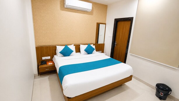 A hotel room with a bed, a nightstand, a trash bin, a wall-mounted air conditioner, and a door. - Hotel Shivaay  (Indore)