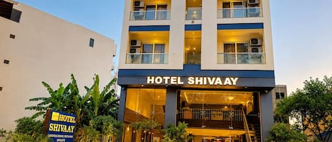 A multi-story hotel with a sign reading 'HOTEL SHIVAAY' and a signboard stating 'HOTEL SHIVAAY LUXURIOUS ROOMS RESTAURANT BANCUETS'.