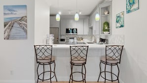 A kitchen with white cabinets, a central island with bar stools, pendant lights, and artwork on the walls. - Golf Cart Included, 3 Pools, Steps to Beach, Pickleball (Santa Rosa Beach)