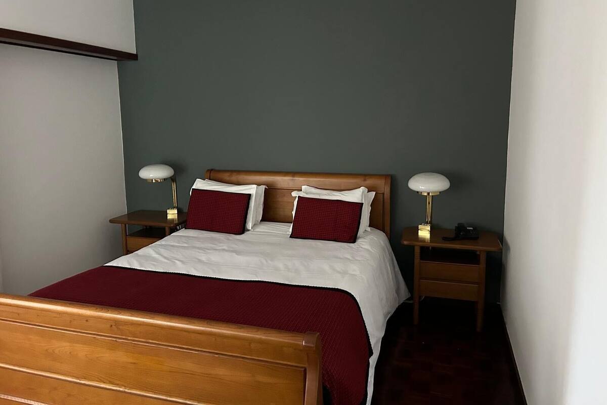Una cama bien hecha con ropa de cama blanca y detalles en rojo, dos mesitas de noche con lámparas y un cabecero de madera.