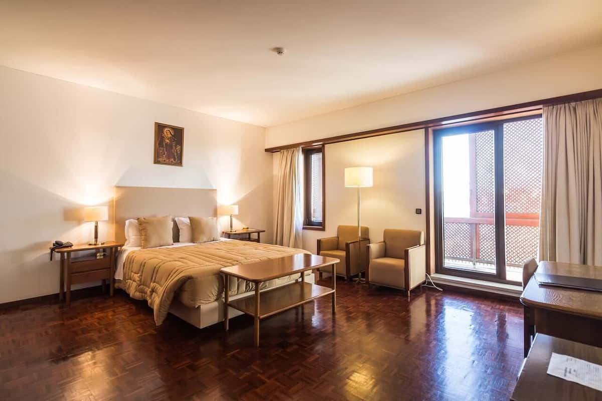 Habitación de hotel con cama, mesitas de noche, escritorio, silla, sofá y balcón con cortinas.