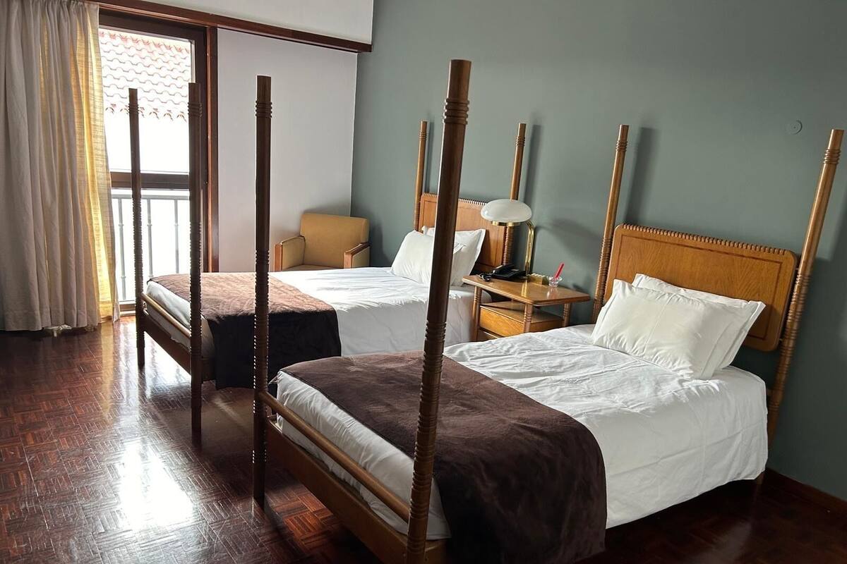 Una cama con dosel y cabecera de madera, una mesita de noche con una lámpara, y una silla en una habitación con piso de madera y vistas a través de una ventana con cortinas.