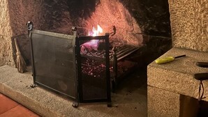 A fireplace with a fire burning inside, a broom, and a yellow tool on a stone ledge. - Lam Hotel Fortaleza de Almeida (Almeida)