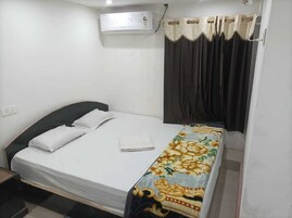 Una cama individual con colchón blanco, colcha estampada y cabecera oscura. Un equipo de aire acondicionado está montado en la pared sobre la cama.