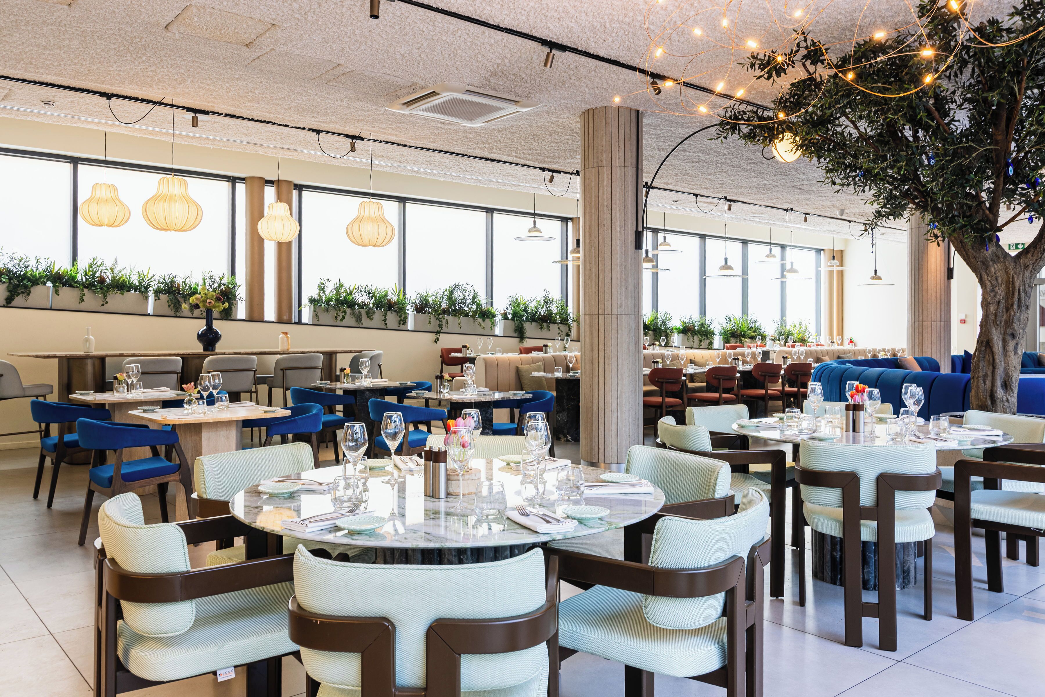 Un restaurante con mesas redondas, sillas, luces colgantes y un árbol grande.